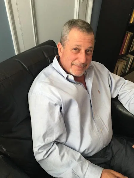 Man with gray hair wearing light blue shirt and dark tie, seated in black chair